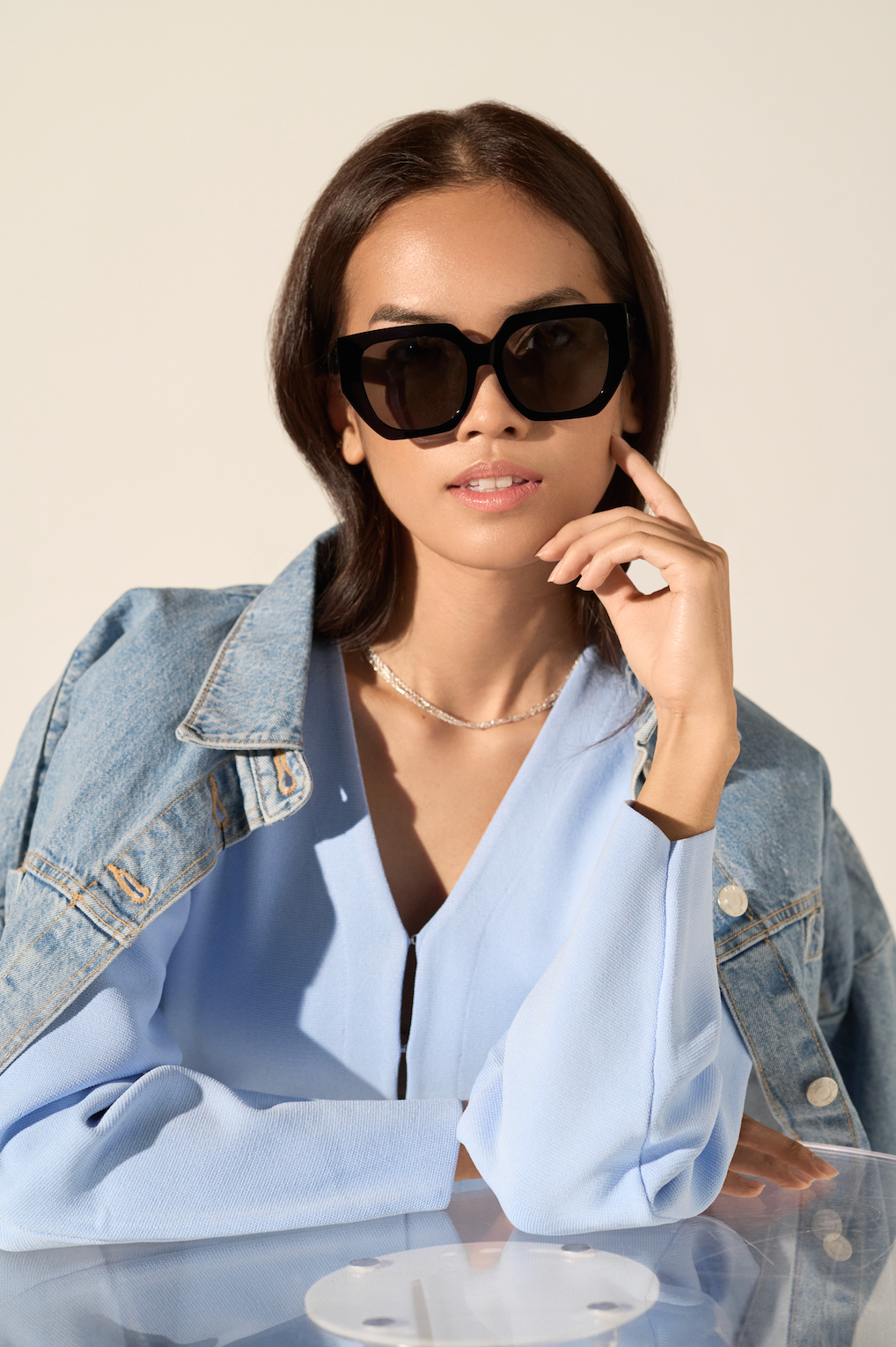 A woman in a light blue top and denim jacket wears KayTran Eyewears Luna - Polished Black sunglasses at a clear table.