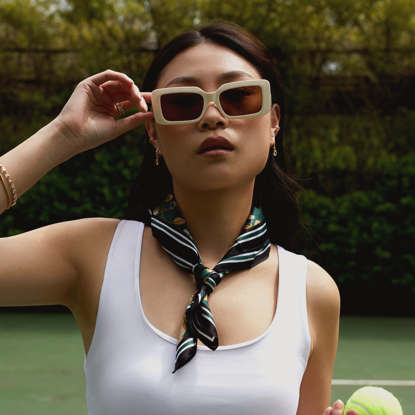 Woman wearing Brooks - Ecru by KayTran Eyewear poses on a tennis court with a scarf and tennis ball, greenery behind her.