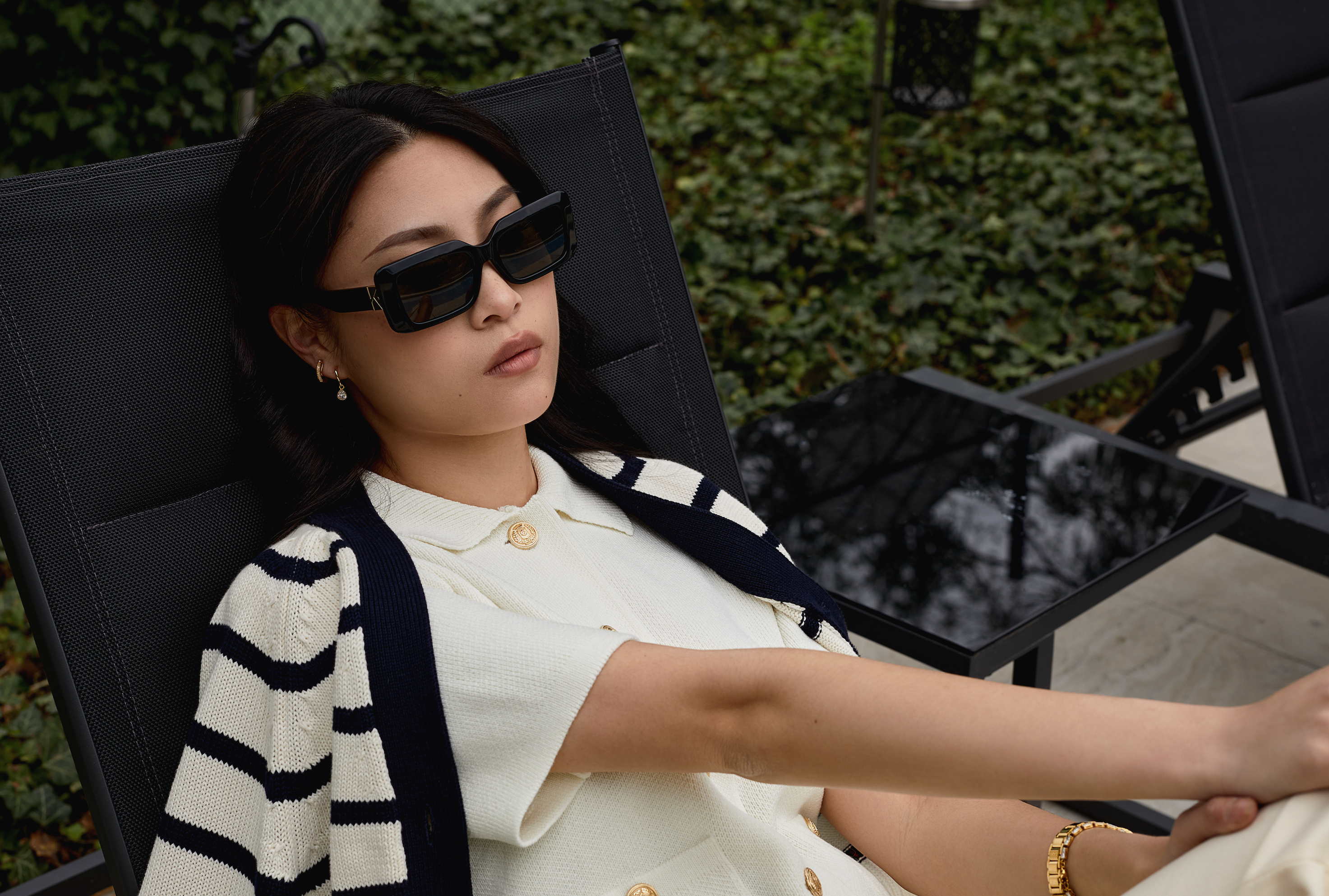 Woman wears KayTran Eyewear Brooks - Polished Black sunglasses, seated outdoors in a white outfit amid patio greenery.