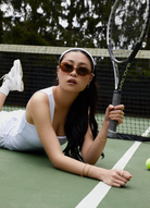 Woman in white athletic wear wears KayTran Eyewears Mara - Latte sunglasses, lying on a tennis court with a racket.