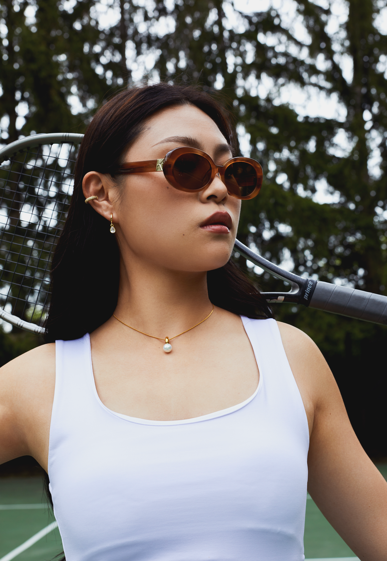 Woman wearing KayTran Eyewear Mara - Latte sunglasses holds a tennis racket on an outdoor court.