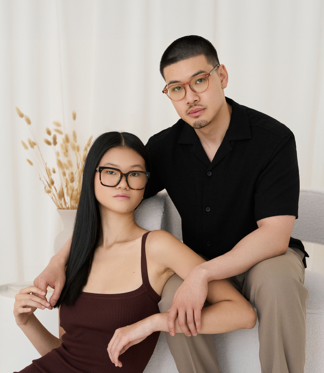 A woman seated and a man standing indoors wear KayTran Eyewears Bailey - Sienna glasses and neutral outfits.