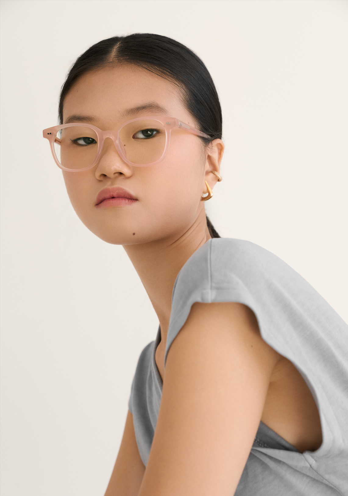 A woman wears KayTran Eyewear’s Easton - Peach Blush pink glasses and a gray shirt, facing the camera plainly.
