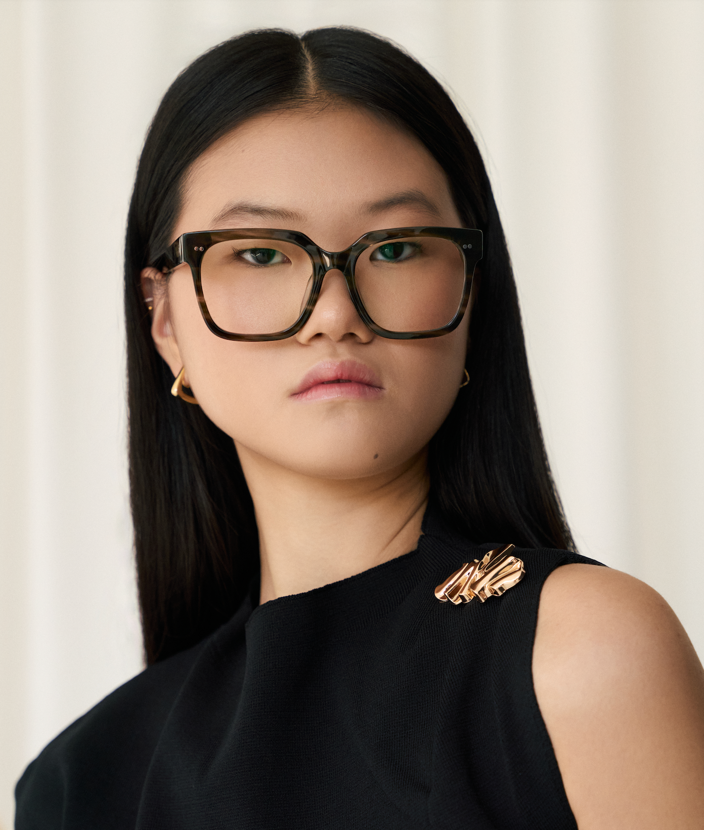 Woman with long black hair wears Andy - Graphite glasses by KayTran Eyewear, gold earrings, and a black sleeveless top.