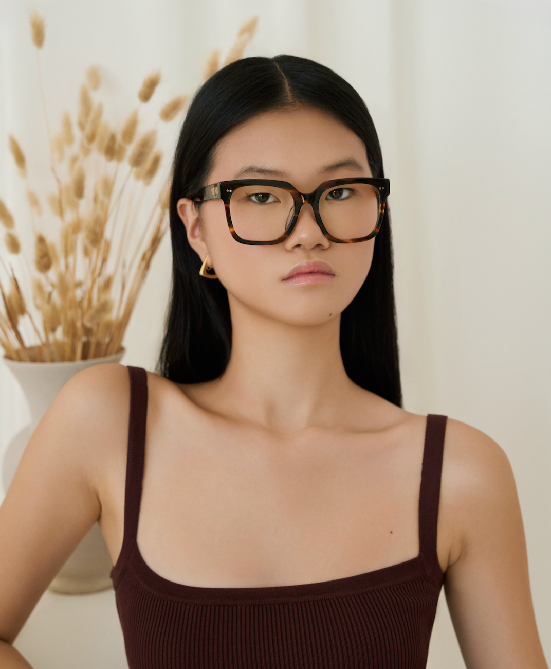 Woman with long black hair wears KayTran Eyewears Andy - Amber Honey glasses and a sleeveless top by a vase of dried grasses.