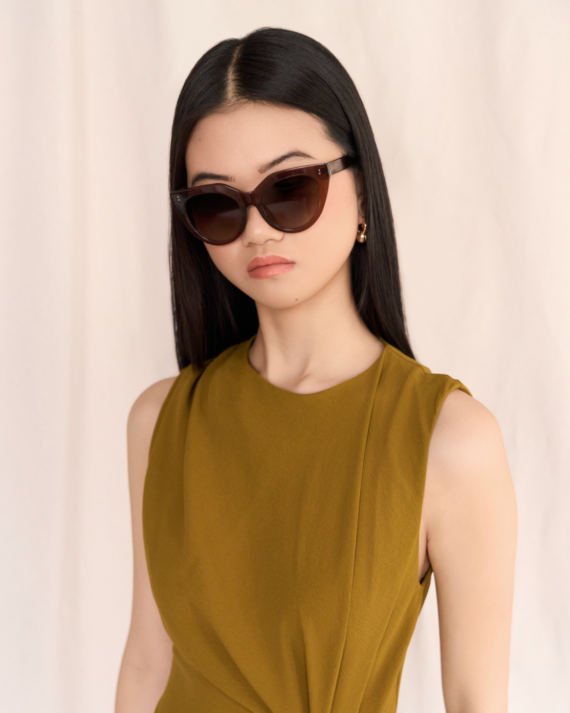Woman with long black hair wears kaytran eyewear Aiden - Truffle sunglasses and a sleeveless olive dress.