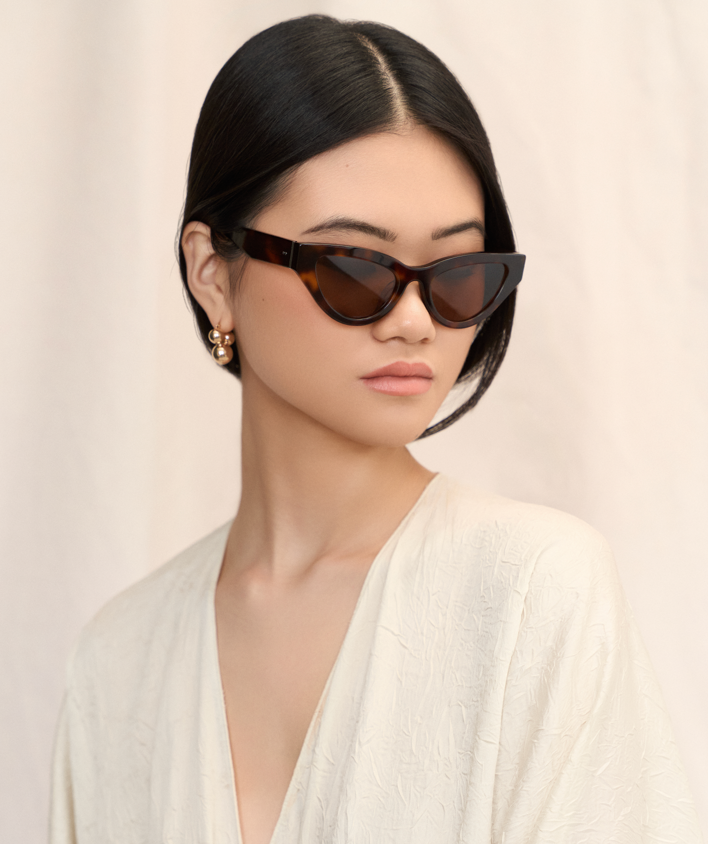 Woman in a cream top wears KayTran Eyewears Drew - Honey Tort cat-eye sunglasses, looking to the side.