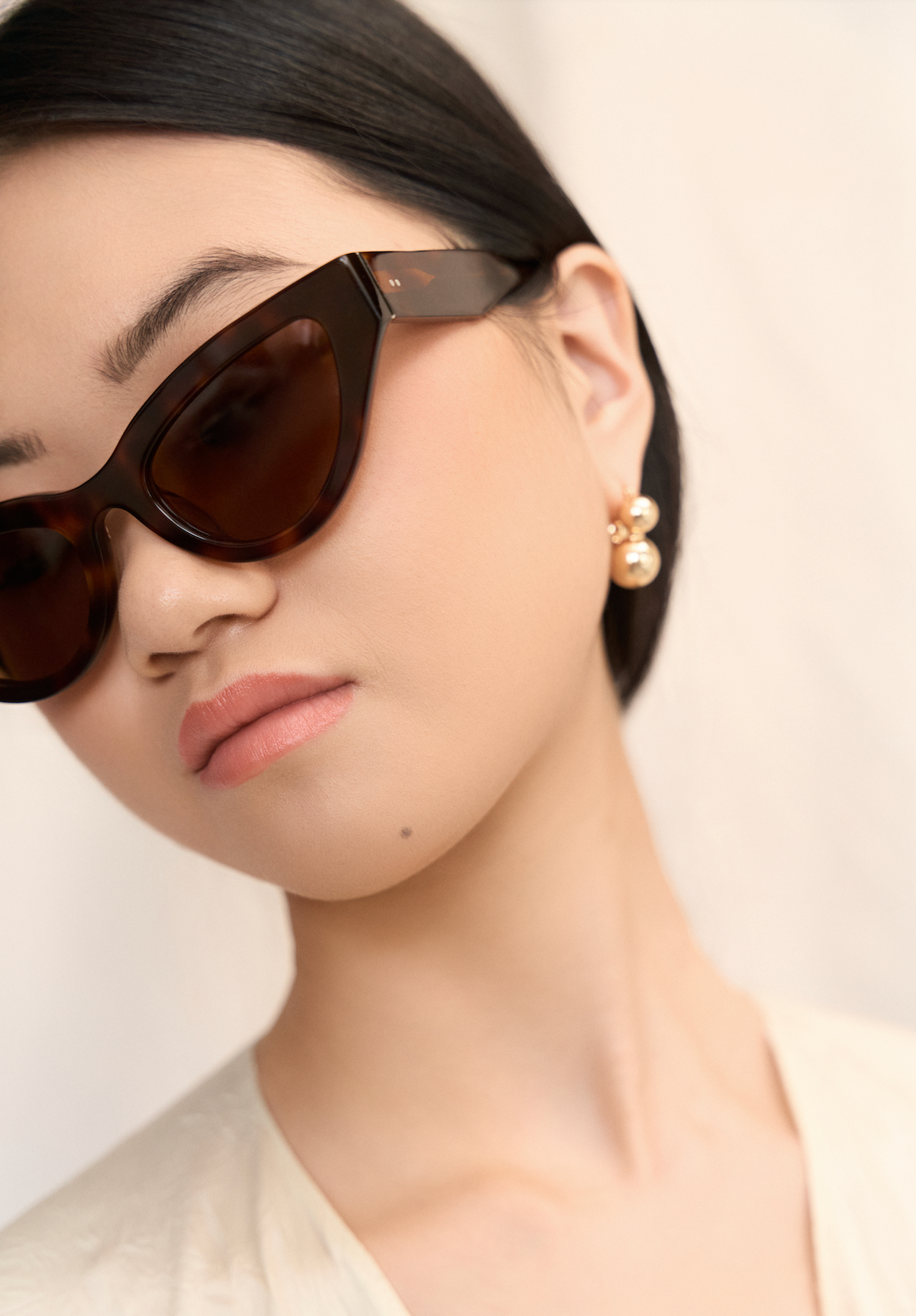 Woman with straight black hair wears KayTran Eyewear Drew - Honey Tort sunglasses and pearl earrings against a neutral backdrop.