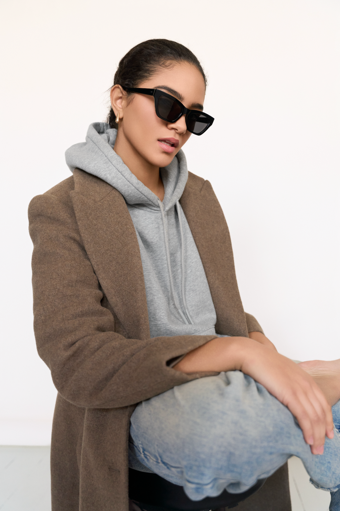 Woman wearing KayTran Eyewears Kai - Polished Black sunglasses, brown coat, gray hoodie, and jeans sits cross-legged.