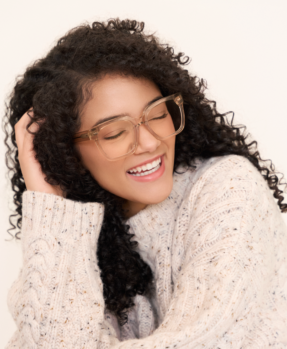 Woman with curly hair smiles with eyes closed, wearing KayTran Eyewear Andy - Folkstone Grey clear frames and a cream sweater.
