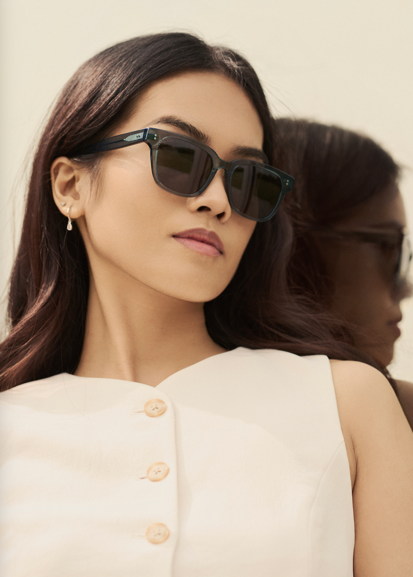 A woman wears KayTran Eyewear’s Charlie - Sage sunglasses and a white top, standing before a mirror.