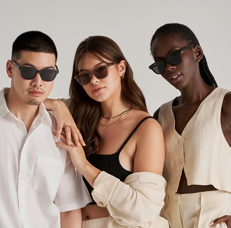 Three people wearing sunglasses and neutral-toned outfits pose together against a plain background.