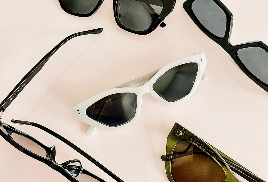 Five pairs of sunglasses with different frame shapes and colors, arranged on a light pink surface.