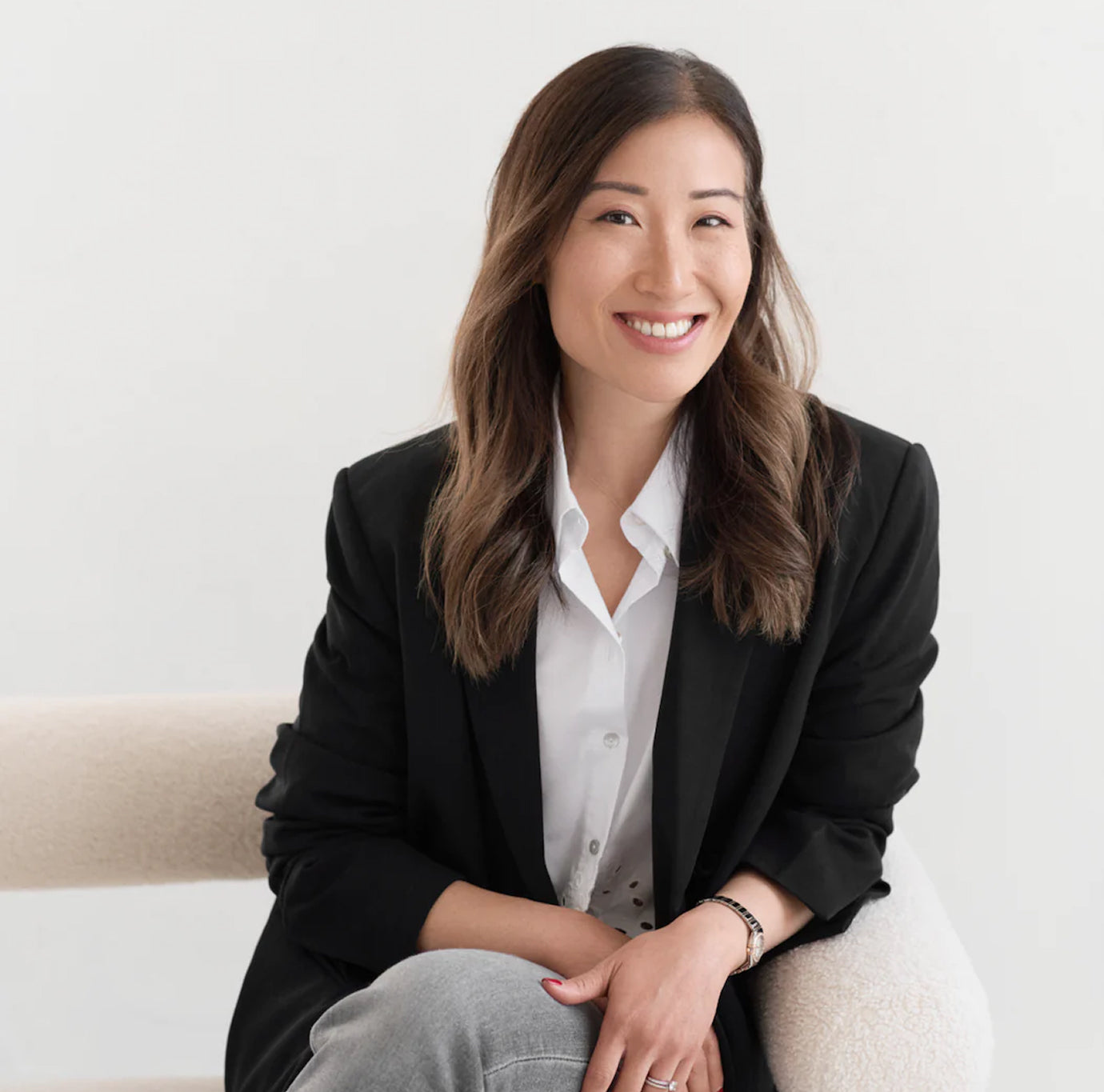 Woman with long brown hair in a black blazer and white shirt seated on a light chair, smiling at the camera.