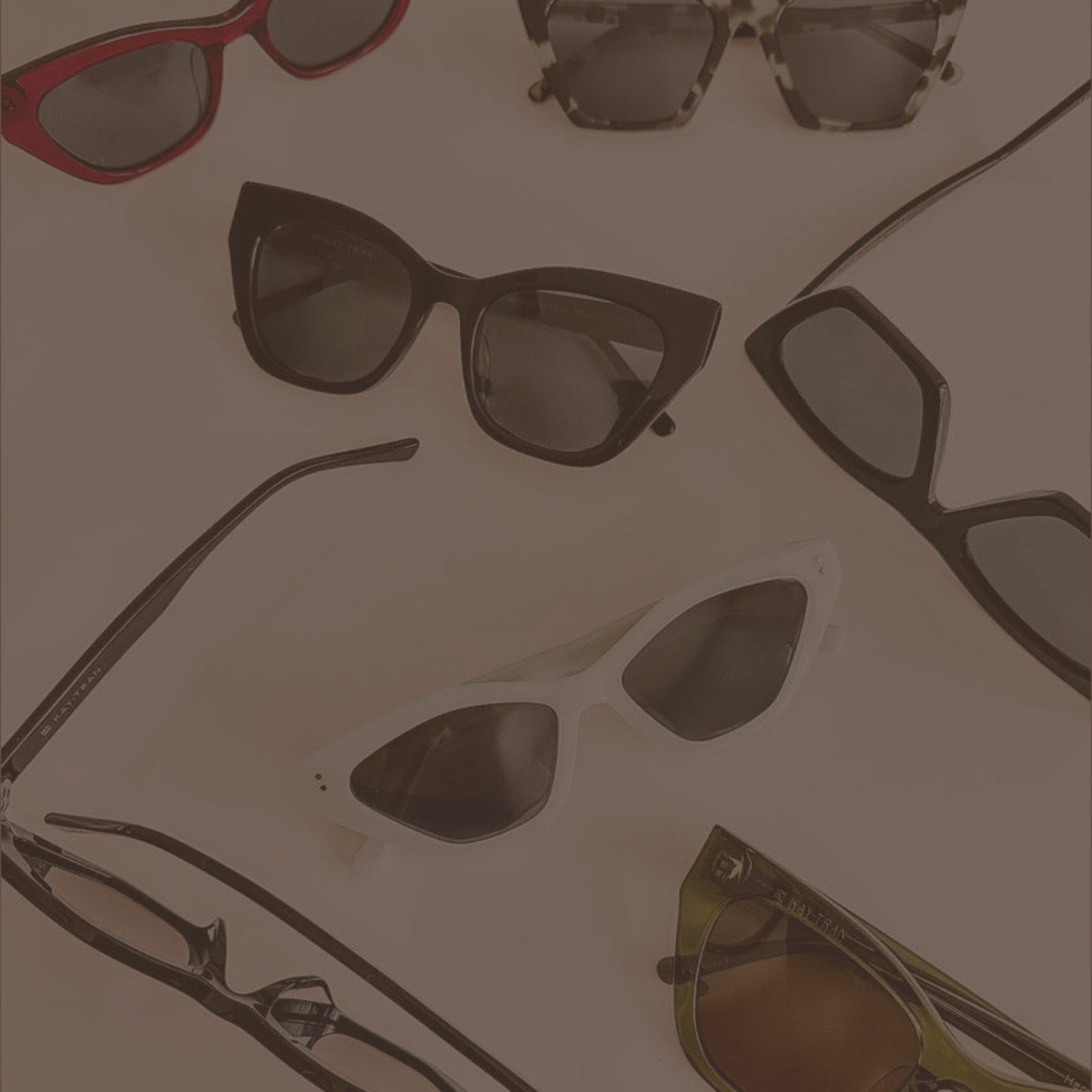 Seven pairs of sunglasses in various shapes and colors are arranged on a flat, neutral-colored surface.