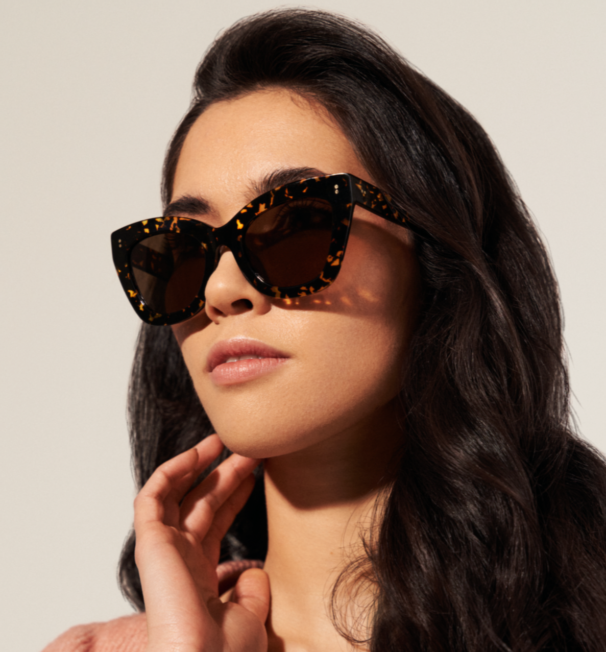 Woman with long dark hair wears KayTran Eyewear Arlo - Amber Scale Tort cat-eye sunglasses and a light pink top.