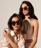 Two women in neutral outfits wear KayTran Eyewears Rowan - Honey Tortoise sunglasses against a plain background.