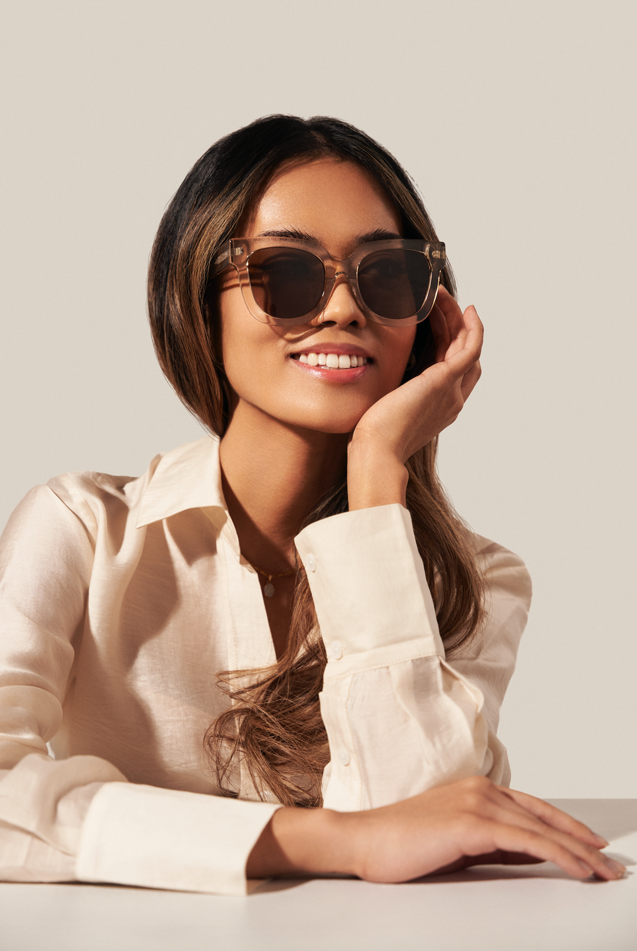 Woman with long hair wears KayTran Eyewear’s Rowan - Sand sunglasses and a cream button-up shirt, smiling at a table.